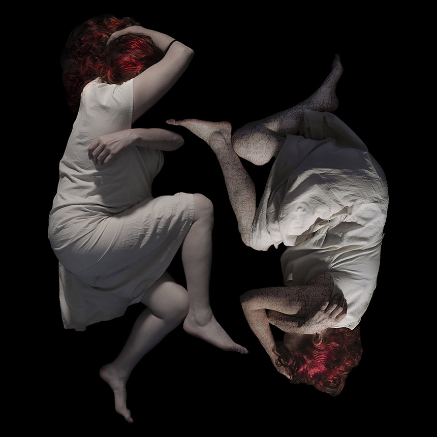 Two female figures dressed in white,  crouched on the ground, facing each other, covering their heads, as if in pain, forming a circle.